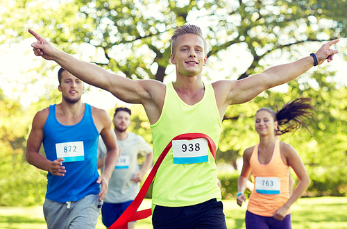 fitness, sport, victory, success and healthy lifestyle concept - happy man winning race and coming first to finish red ribbon over group of sportsmen running marathon with badge numbers outdoors