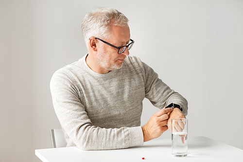 medicine, healthcare and people concept - senior man with glass of water water and pill looking at wristwatch or smartwatch with health application at home