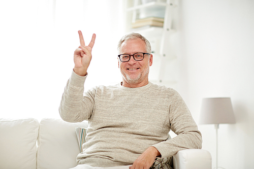 old age, gesture and people concept - smiling senior man in glasses sitting on sofa and showing v sign at home