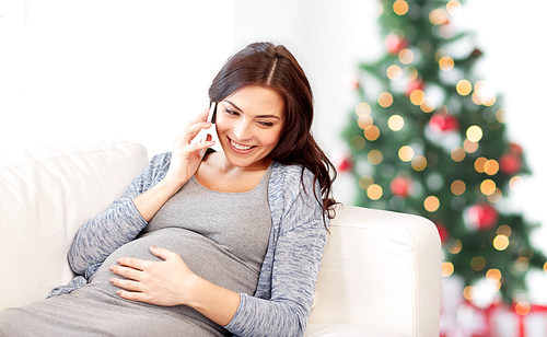 pregnancy, holidays, technology, people and expectation concept - happy pregnant woman calling on smartphone on couch over christmas tree background
