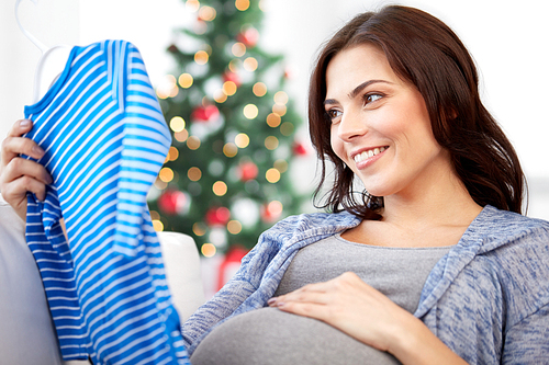 holidays, pregnancy, people and kids clothing concept - happy woman holding and looking at blue baby boys bodysuit over christmas tree background