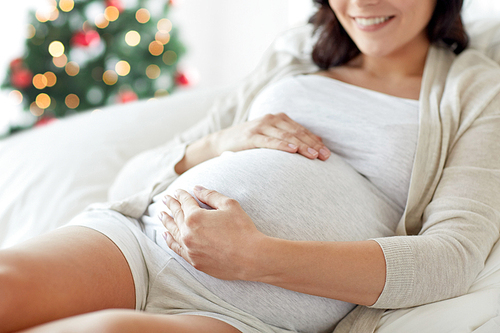 pregnancy, rest, people and expectation concept - close up of happy smiling pregnant woman lying in bed and touching her belly over christmas tree background