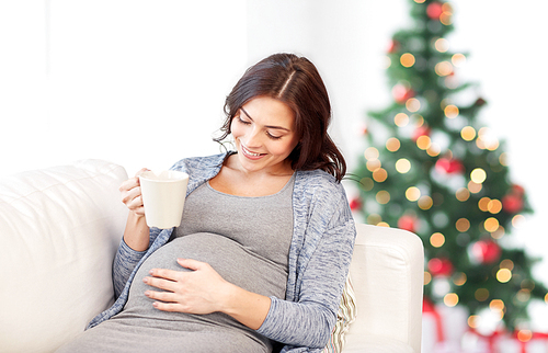 pregnancy, holidays, people and expectation concept - happy pregnant woman with cup drinking tea on sofa over christmas tree background