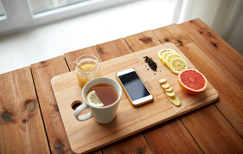 health, traditional medicine, folk remedy and ethnoscience concept - smartphone with cup of ginger tea, honey and citrus on wooden board