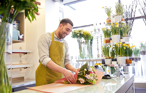 people, shopping, sale, floristry and consumerism concept - happy smiling florist man wrapping flowers in paper at flower shop