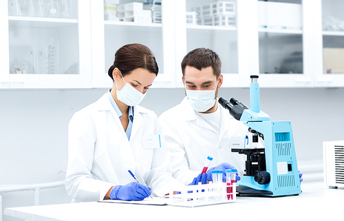 science, chemistry, technology, biology and people concept - young scientists with test tube and microscope making research in clinical laboratory and taking notes