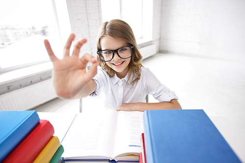 education, vision, elementary school, learning and people concept - happy smiling girl in eyeglasses with books sitting at table and showing ok sign