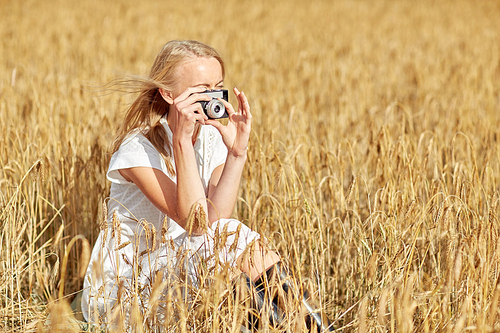 nature, summer holidays, vacation and people concept - happy young woman taking picture with vintage film camera in cereal field