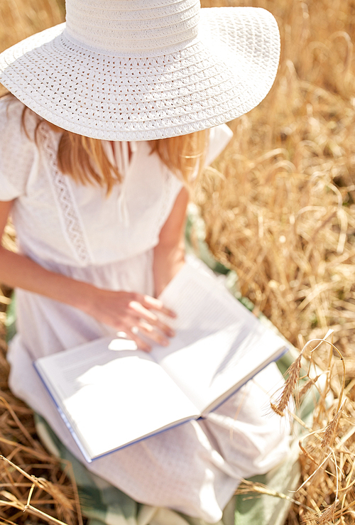 country, summer holidays, literature and people concept - close up of young woman in white straw hat and dress reading book on cereal field