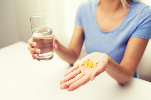 healthy eating, medicine, health care, food supplements and people concept - close up of woman hands holding pills or fish oil capsules and water glass at home