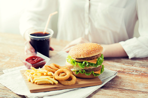 fast food, people and unhealthy eating concept - close up of woman hands with hamburger or cheeseburger, french fries, squid rings and cola drink sitting at table