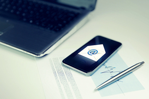 business, technology and communication concept - close up of smartphone with email message icon , laptop computer and chart with pen on office table