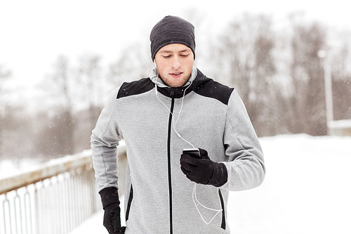 fitness, sport, people, technology and healthy lifestyle concept - young man in earphones with smartphone listening to music and running along winter road