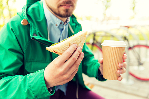 people, junk food, eating and lifestyle - close up of young man with coffee cup and sandwich eating and drinking on city street
