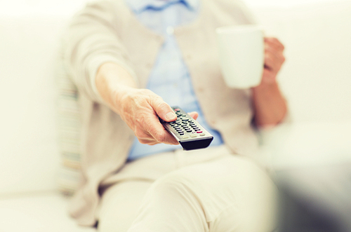technology, television, age and people concept - close up of senior woman watching tv, drinking tea and changing channels by remote control at home