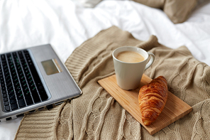 coziness, morning, technology and breakfast concept - bedroom with laptop computer, coffee cup and croissant on bed at home