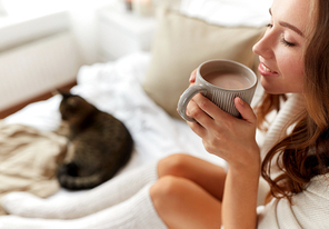 winter, coziness, leisure and people concept - close up of happy young woman with cup of coffee or cocoa drink and cat in bed at home