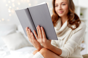 christmas, comfort, leisure and people concept - happy young woman reading book in bed at home bedroom