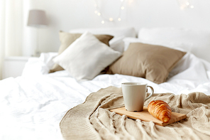 breakfast, coziness, morning, holidays and winter concept - cozy bedroom with coffee cup and croissant on wooden board in bed at home