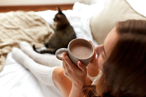 winter, coziness, leisure and people concept - close up of happy young woman with cup of coffee or cacao and cat in bed at home