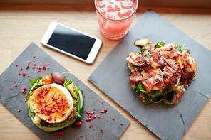 food, eating and technology concept - goat cheese and prosciutto ham salads on stone plates with smartphone and glass of drink at restaurant or cafe