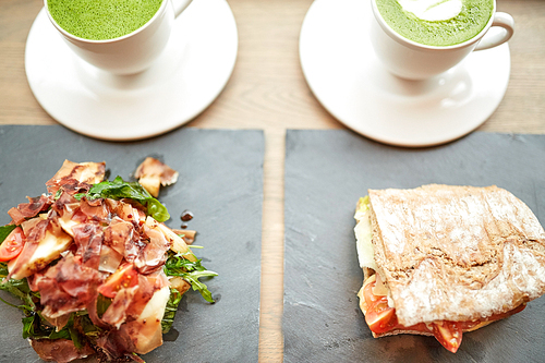 food, dinner, haute cuisine and eating concept - prosciutto ham salad with panini sandwich on stone plates and cups of matcha green tea latte at restaurant