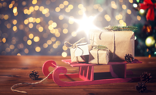 holidays, presents, new year and celebration concept - close up of gift boxes with blank note on red wooden sleigh over christmas tree and lights background