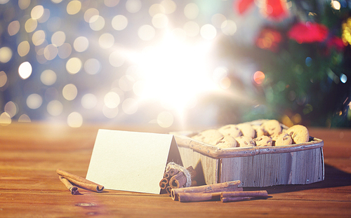 christmas, holidays, food and baking concept - close up of oat cookies in wooden box with blank greeting card and cinnamon on table over lights