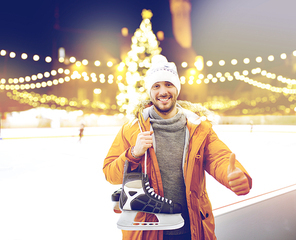 christmas and people concept - happy young man with ice-skates showing thumbs up on skating rink over outdoor holiday lights background
