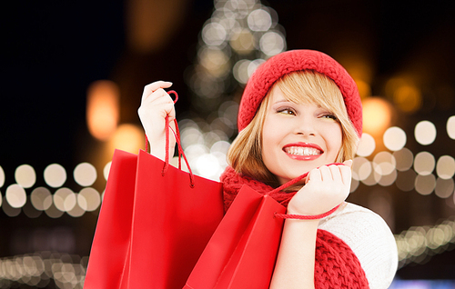 winter holidays, sale and people concept - smiling young woman in hat and scarf with red shopping bags over christmas tree lights background