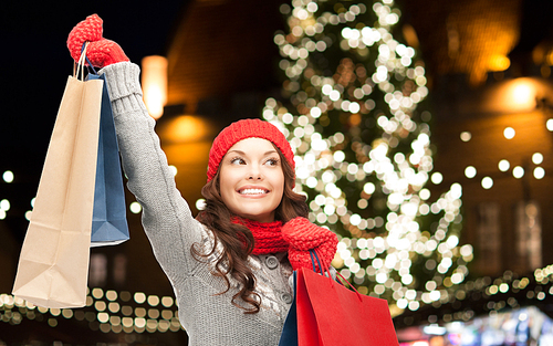 winter holidays, sale and people concept - smiling young woman in hat and scarf with shopping bags over christmas tree lights background