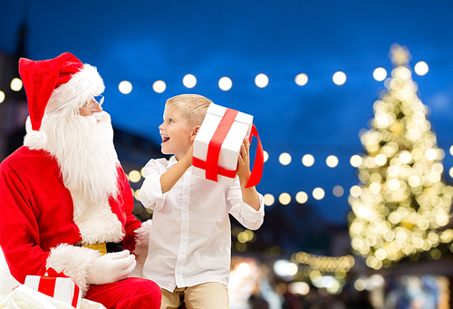 holidays and people concept - santa claus and happy little boy with gift box over christmas tree lights background