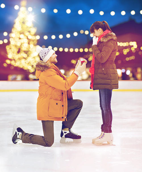 love, holidays and relationships concept - happy couple with engagement ring at outdoor skating rink over christmas lights background