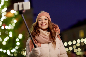 holidays and people concept - beautiful happy young woman taking picture by selfie stick over christmas tree lights in winter evening