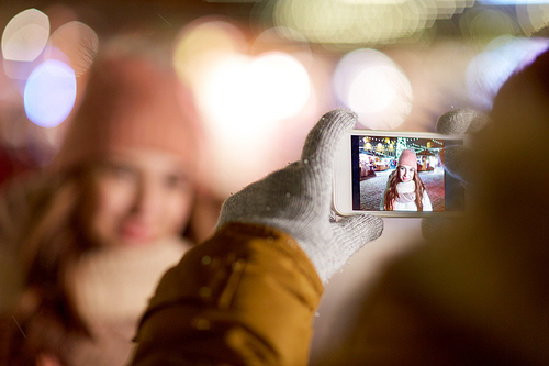 holidays, winter and people concept - hand with smartphone taking picture of young woman at christmas street market