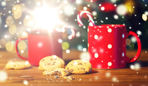 holidays, christmas, winter, food and drinks concept - close up of candy canes and cups on wooden table over lights