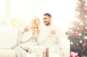 family, christmas, holidays, love and people concept - happy couple covered with plaid drinking tea and sitting on sofa at home