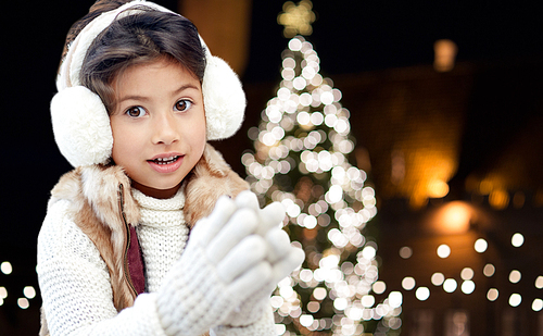winter, holidays and people concept - happy little girl wearing earmuffs over christmas tree lights background