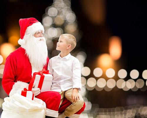 holidays and people concept - santa claus and happy little boy with gift box over christmas tree lights background