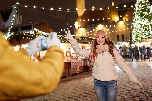 holidays, winter and people concept - happy woman posing for boyfriend with smartphone on square at christmas market