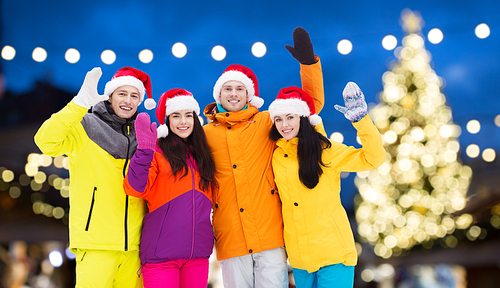 winter holidays, friendship and people concept - happy friends in santa hats and ski suits waving hands outdoors over christmas lights background