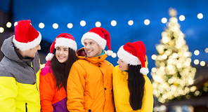 winter holidays, friendship and people concept - happy friends in santa hats and ski suits outdoors over christmas lights background