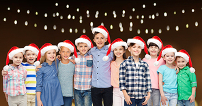 childhood, holidays, christmas and people concept - happy smiling children in santa hats hugging over lights background