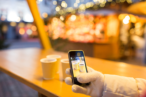 winter holidays, technology and people concept - hand with mulled wine or hot chocolate drinks picture on smartphone at christmas market