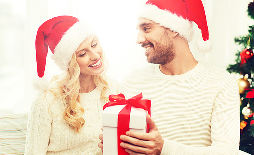 christmas, holidays and people concept - happy couple in santa hats with gift box sitting on sofa at home