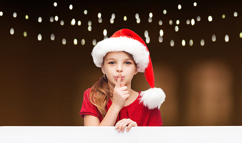 christmas, holidays and advertisement concept - happy girl child in santa helper hat with blank white board making hush gesture over garland lights background