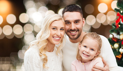 family, holidays and people concept - happy mother, father and little daughter sitting on sofa at christmas tree over lights background