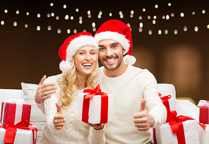 christmas, holidays and people concept - happy couple in santa hats with gift boxes sitting on sofa and showing thumbs up over garland lights background