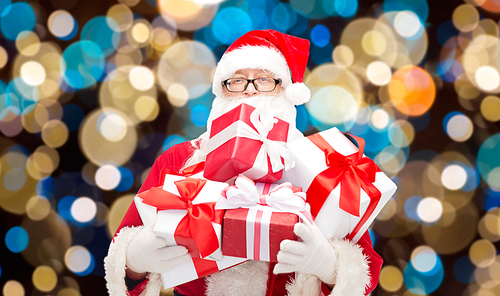 christmas, holidays and people concept - man in costume of santa claus with gift boxes over lights background