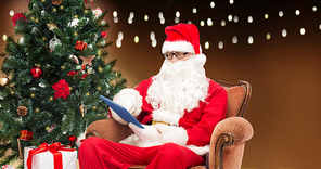 technology, holidays and people concept - man in costume of santa claus with tablet pc computer, gift and christmas tree sitting in armchair over garland lights background